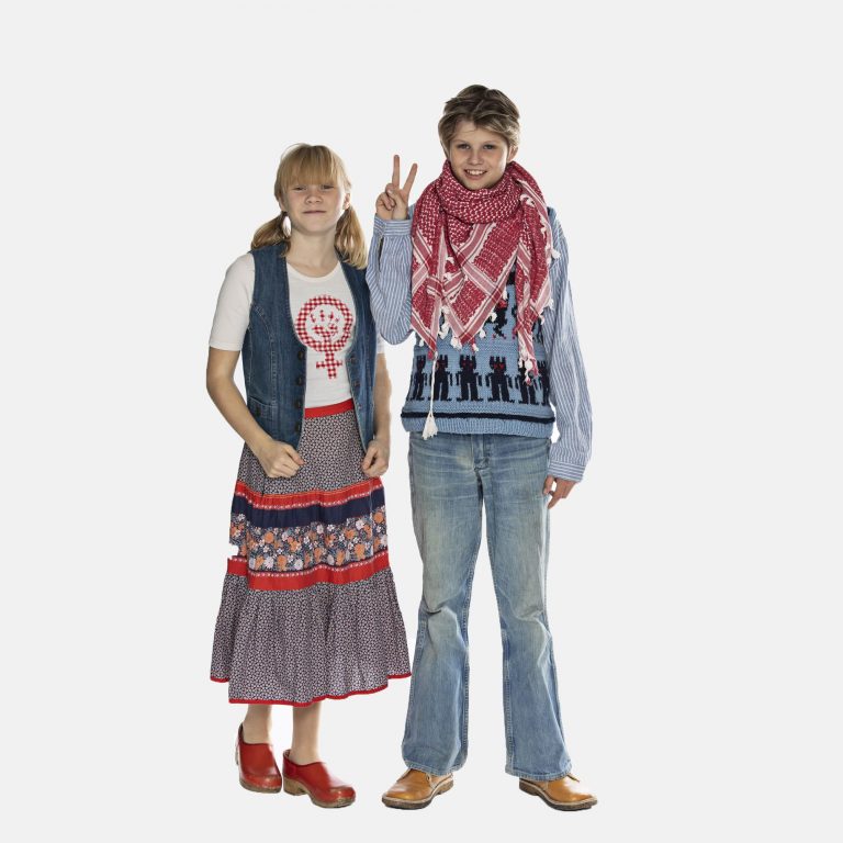 Dreng fra 1970'erne med hjemmestrikket sweater, hvidt og rødt partisanertørklæde, vidde cowboybukser, fodformede brune sko og peace-tegn, med 70'er-pige i lang nederdel i farver og mønstre, peace-t-shirt, cowboyvest, røde træsko og rottehaler, læs om moden i 1970'erne, originalt tøj fra museet Tidens Samling, museum for klæder, form og bolig i det 20. århundrede, Kulturmaskinen, Farvergården, Brandts Klædefabrik, Odense